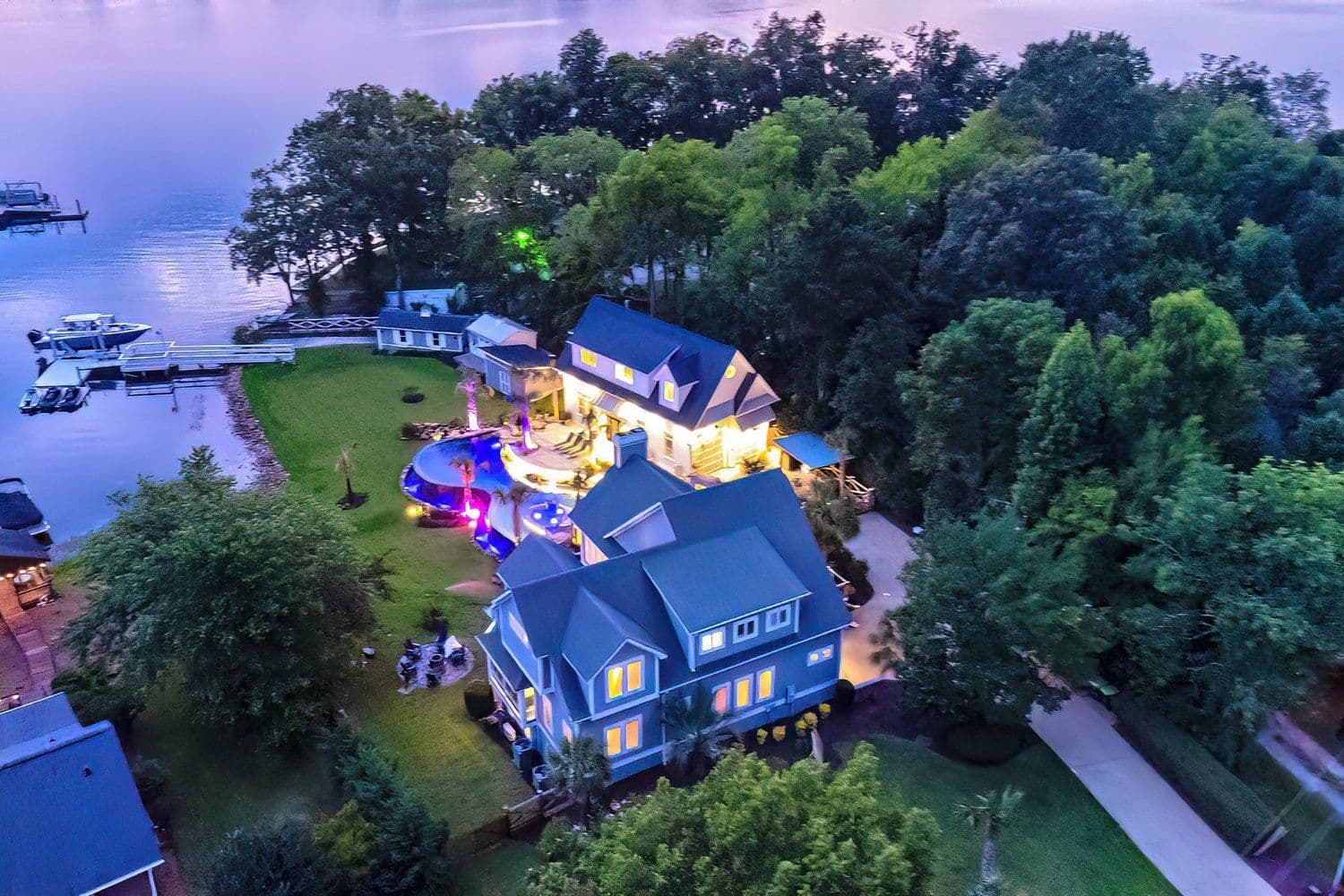 Dusk aerial of a lakefront estate with colorful pool lighting