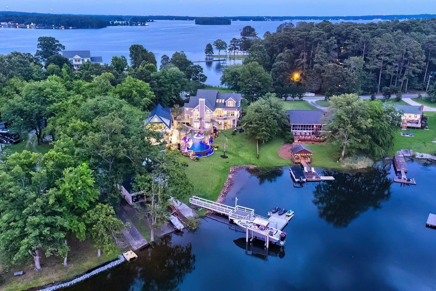Dusk aerial of a lit lakefront house with several boat docks among trees